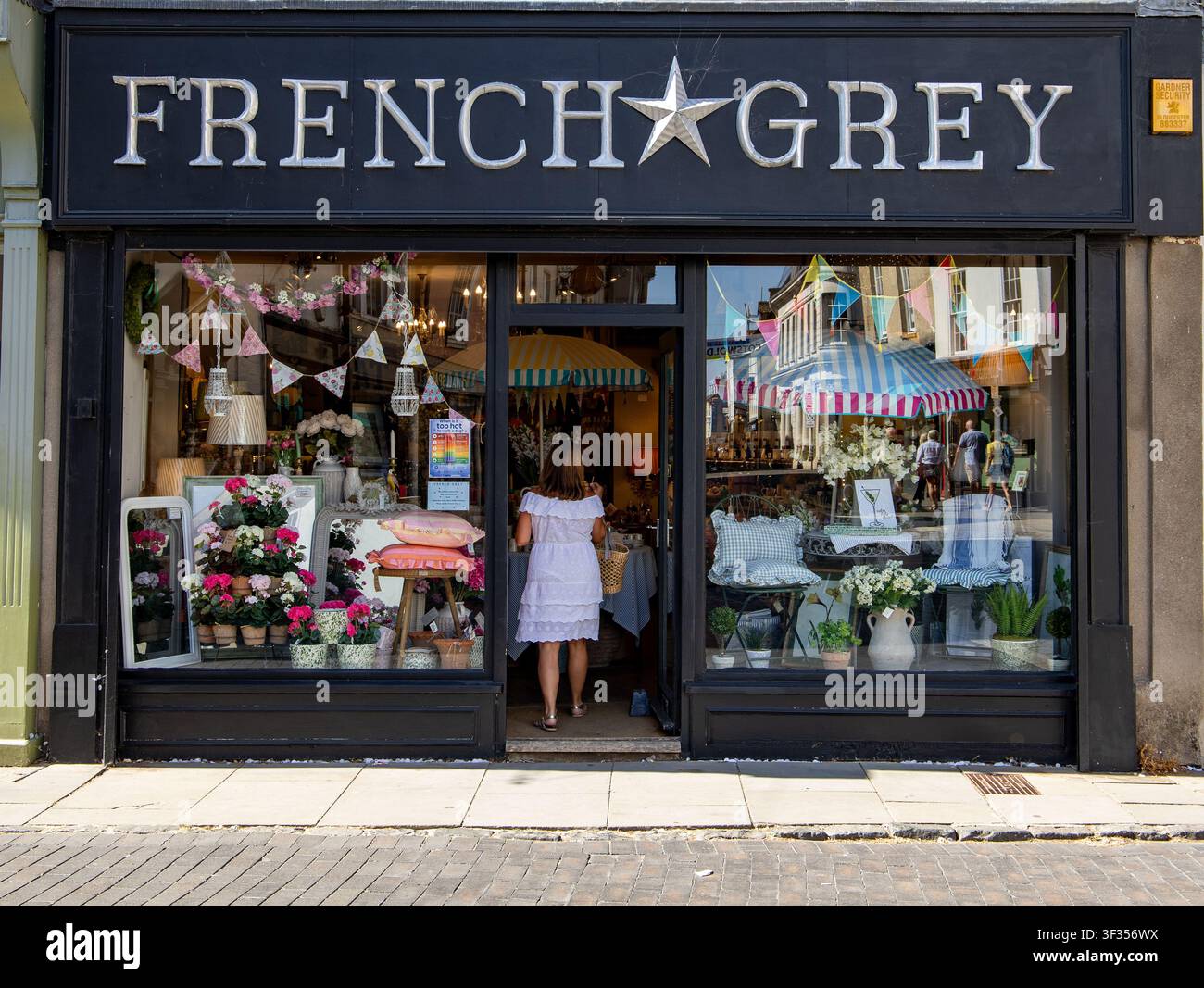 Donna in abito bianco che entra in un negozio boutique French Grey con vetrina decorata piena di fiori e oggetti per la casa a Cirencester, UK Foto Stock