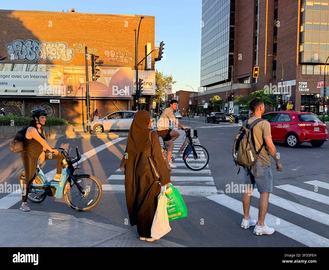 Pedoni e ciclisti multiculturali attendono a un passaggio pedonale su Jean-Talon Street a Montreal, Quebec, Canada, alla luce calda della sera Foto Stock