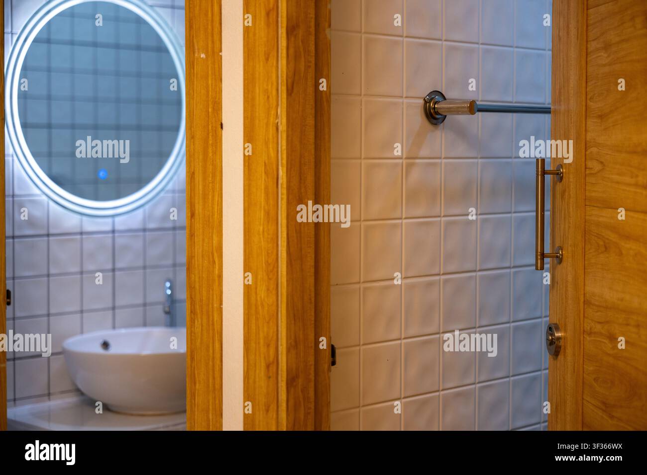 Un interno di bagno moderno con specchio rotondo illuminato e lavandino in ceramica visibili attraverso una porta di legno, le rifiniture in legno caldo e il muro piastrellato creano un'atmosfera accogliente Foto Stock
