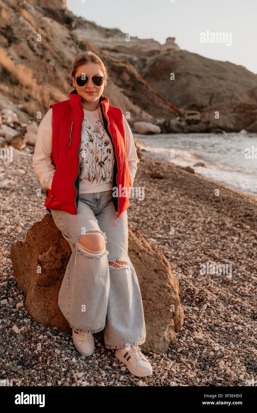 Donna spiaggia costa Giovane donna sorridente con un giubbotto rosso e jeans strappati seduta su una roccia in riva al mare al tramonto Foto Stock