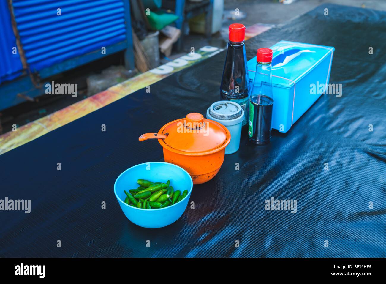 Una varietà di condimenti da tavolo tra cui peperoni verdi in una ciotola blu, contenitore per salsa arancione, bottiglie di salsa di soia e una scatola di fazzoletti posizionati su un bl Foto Stock