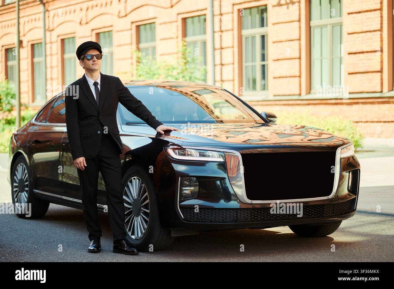 Scatto a tutta lunghezza di un giovane autista caucasico in piedi accanto a una berlina di lusso, in attesa del passeggero su una strada cittadina. Ideale per trasporti premium, viaggi aziendali, marketing di concierge Foto Stock