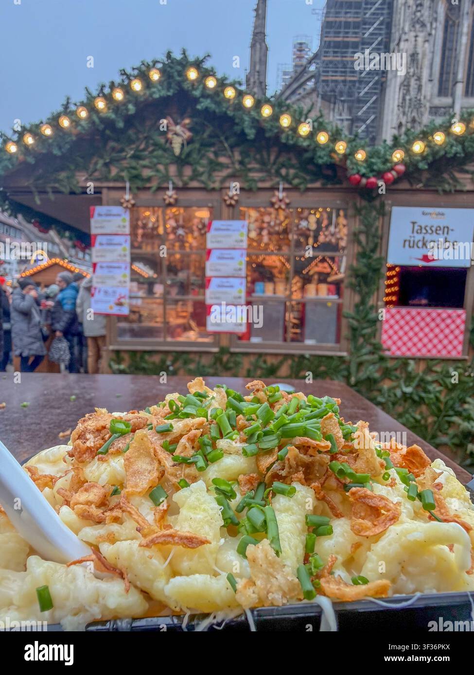 ULM, GERMANIA - 13 DICEMBRE 2025: Primo piano di tradizionali Kässpätzle svevi con cipolle fritte e erba cipollina, serviti al mercato natalizio di Ulma. Foto Stock