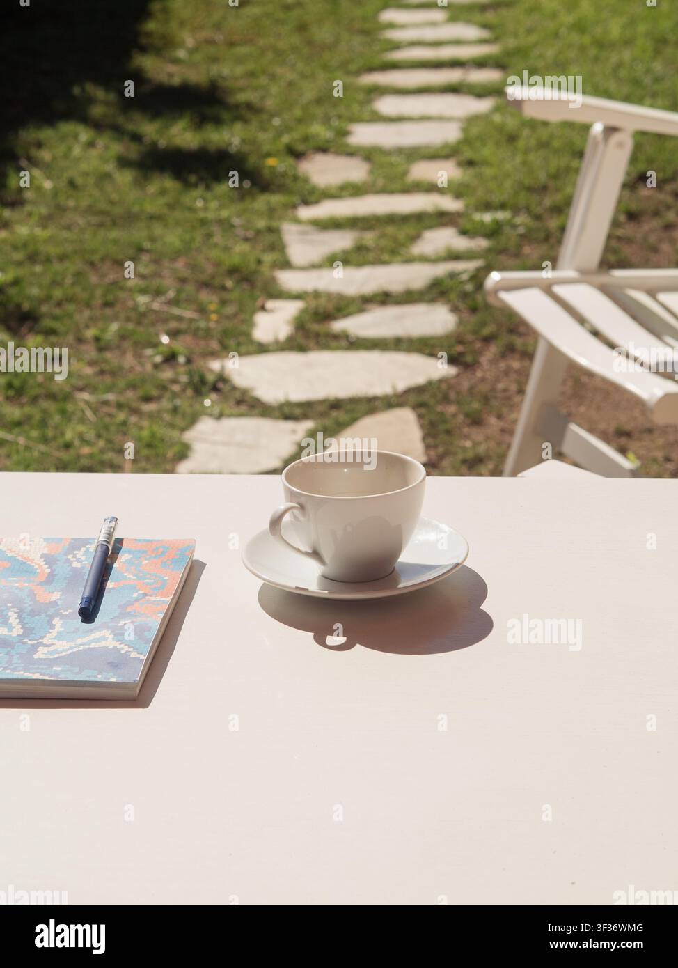 Tavolo da esterno bianco con caffè, quaderno e penna accanto a un sentiero di pietra e una sdraio sull'erba verde, vista verticale dall'alto Foto Stock