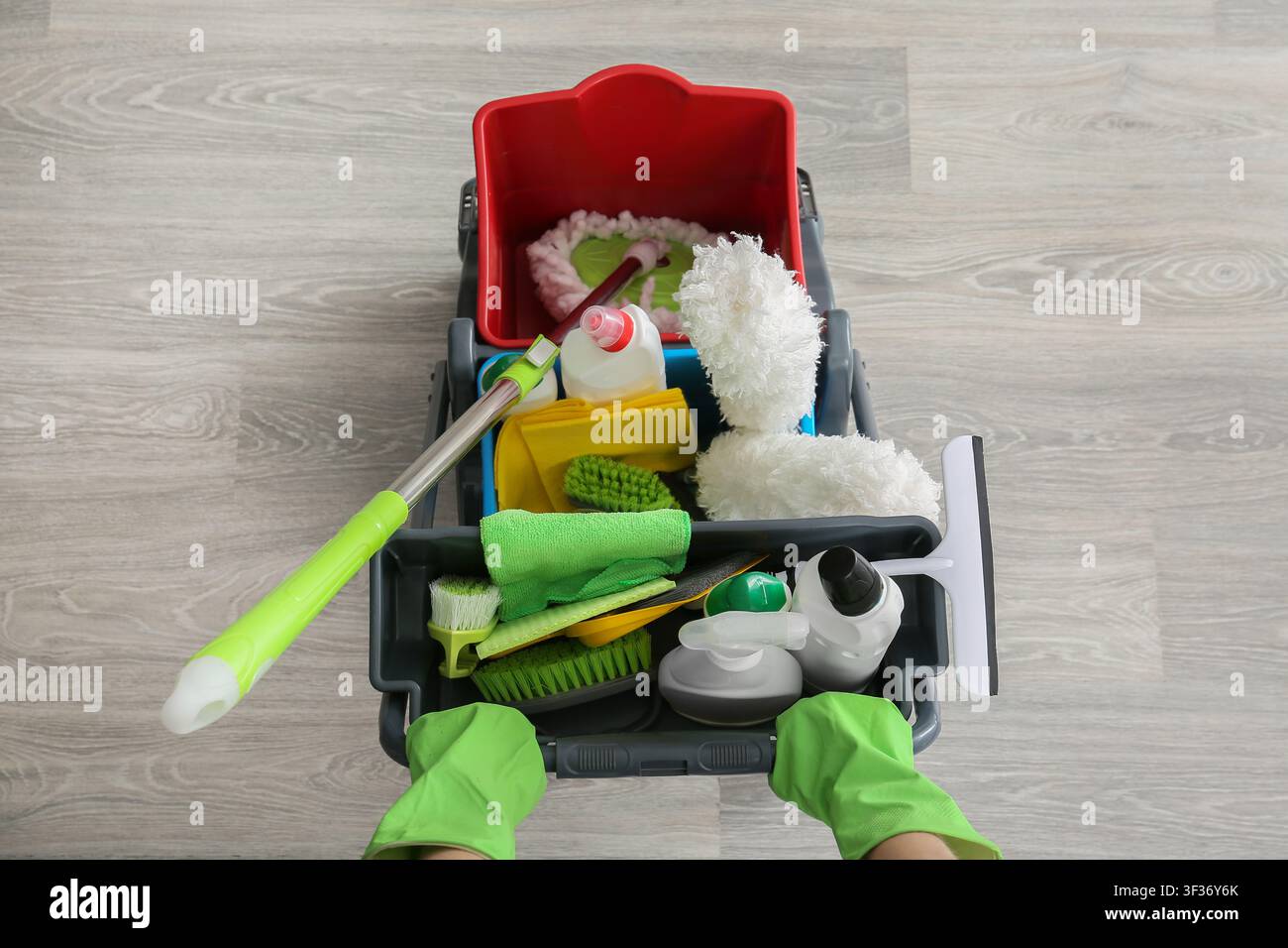 Persona con guanti verdi che spinge un carrello per la pulizia pieno di vari prodotti e strumenti per la pulizia della casa Foto Stock