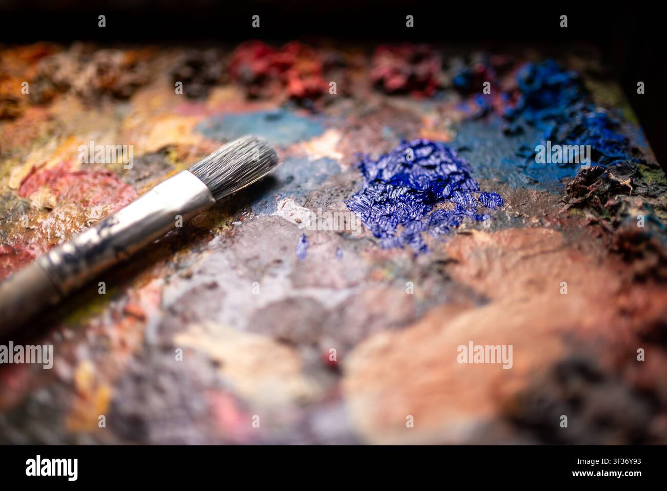 Primo piano di un pennello usato appoggiato su una tavolozza artistica ruvida con pitture a olio miste e colori vivaci Foto Stock
