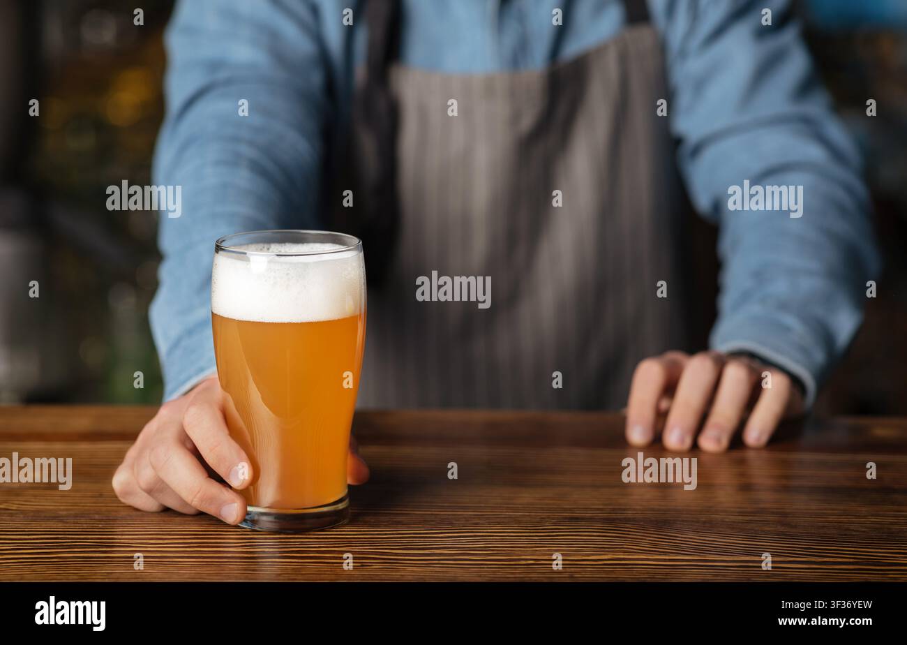 Pinta di birra artigianale. Barman in grembiule e camicia blu messo su vetro di tavola di legno di birra chiara Foto Stock