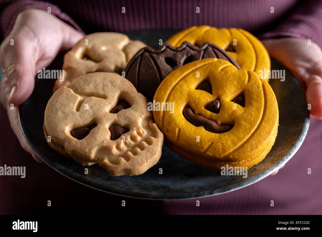 Primo piano di biscotti di Halloween fatti in casa a forma di zucche, teschi, pipistrelli e fantasmi serviti su un piatto scuro tenuto da una donna. Foto Stock