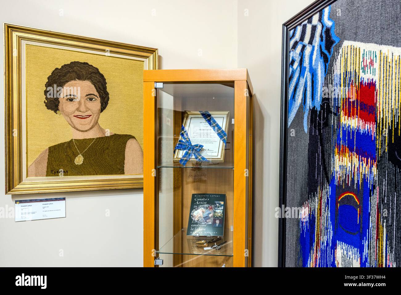 Un arazzo ritratto e una vetrina con un premio e una pubblicazione sull’artista tessile Elizabeth Lefort (1914–2005) nella galleria a lei dedicata al Centro Culturale Les Trois Pignons sul Cabot Trail a Cheticamp, nella Nuova Scozia, Canada. Lefort è considerata una delle artiste di tappeti uncinati più conosciute al mondo; le sue opere sono esposte a Buckingham Palace, alla Casa Bianca e al Vaticano, tra gli altri luoghi. Cheticamp, Nuova Scozia, Canada Foto Stock