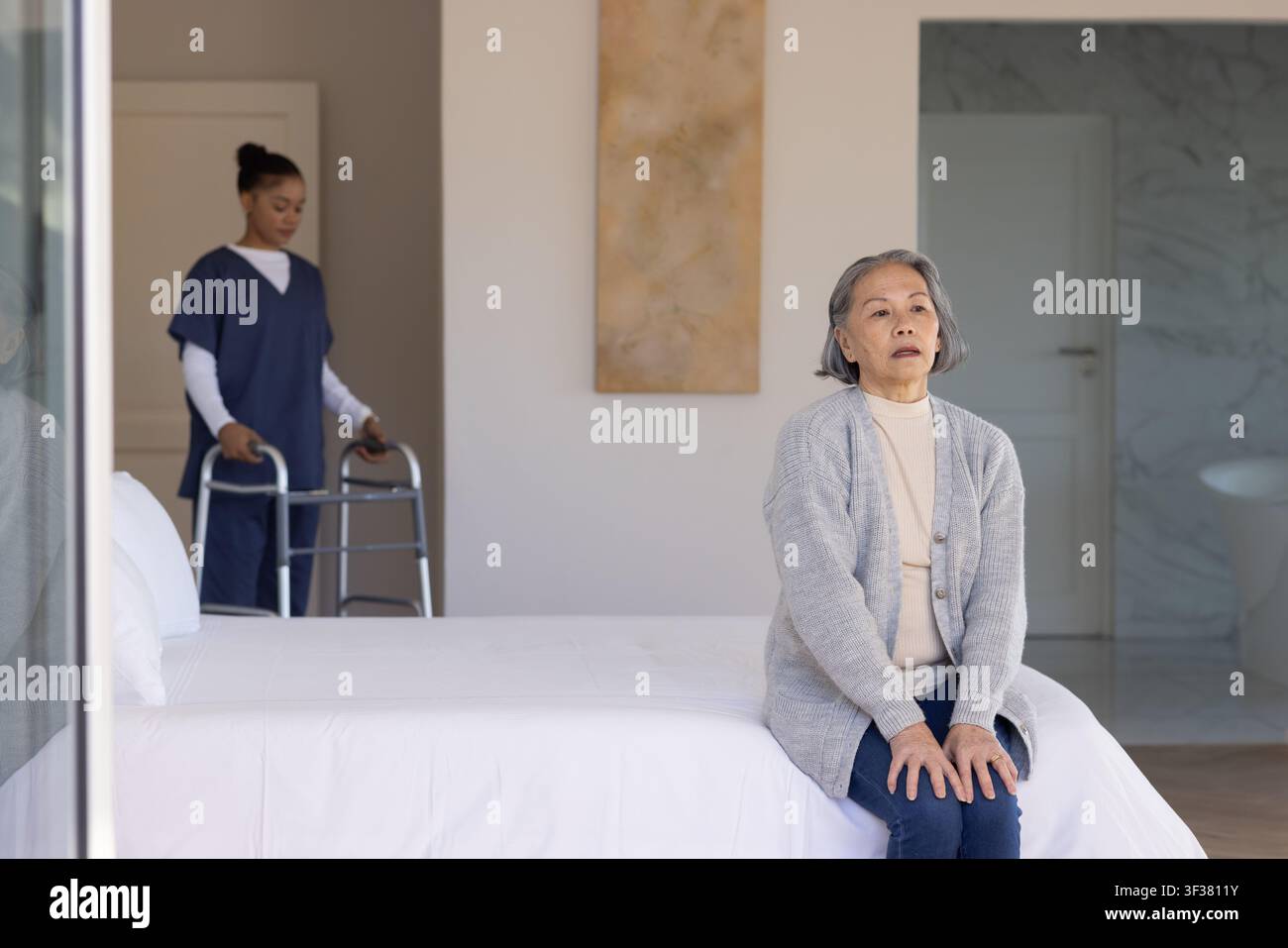 Donna anziana asiatica seduta sul letto, donna afroamericana badante che tiene un deambulatore, spazio di copia Foto Stock