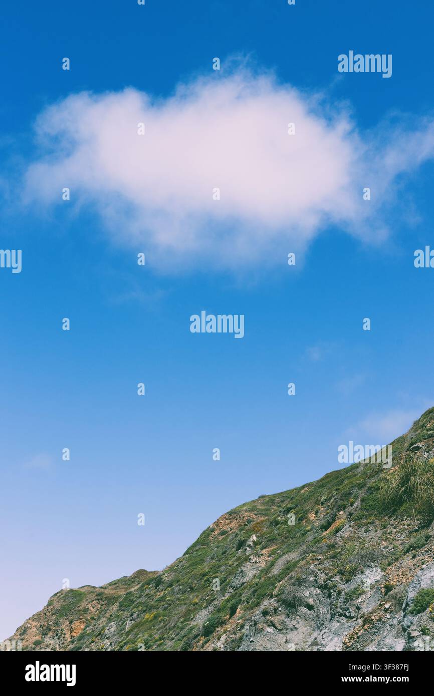 Una nuvola bianca e soffice in un cielo blu brillante con una sezione di montagna in primo piano. Foto Stock