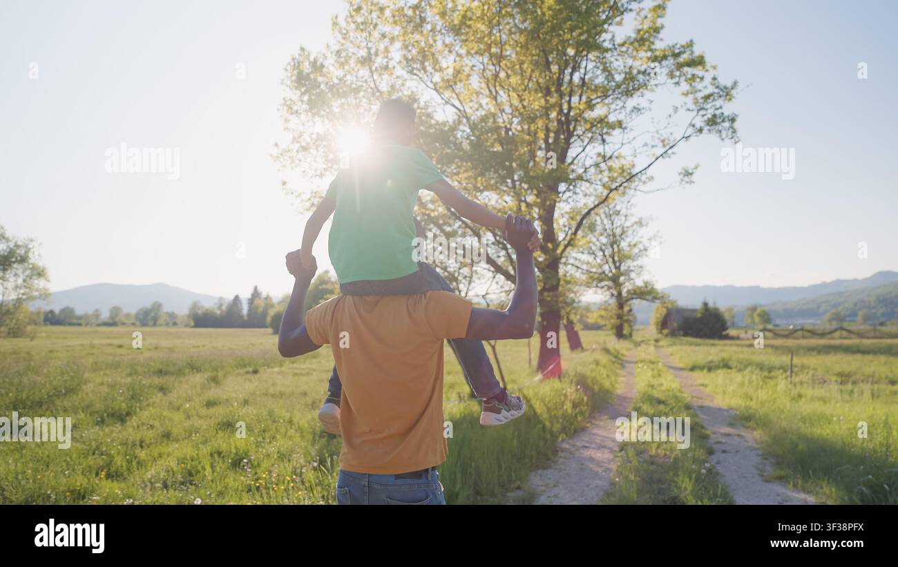 Un padre porta il suo figlio piccolo sulle spalle in un campo al tramonto. Condividono risate e creano ricordi nella natura. Foto Stock