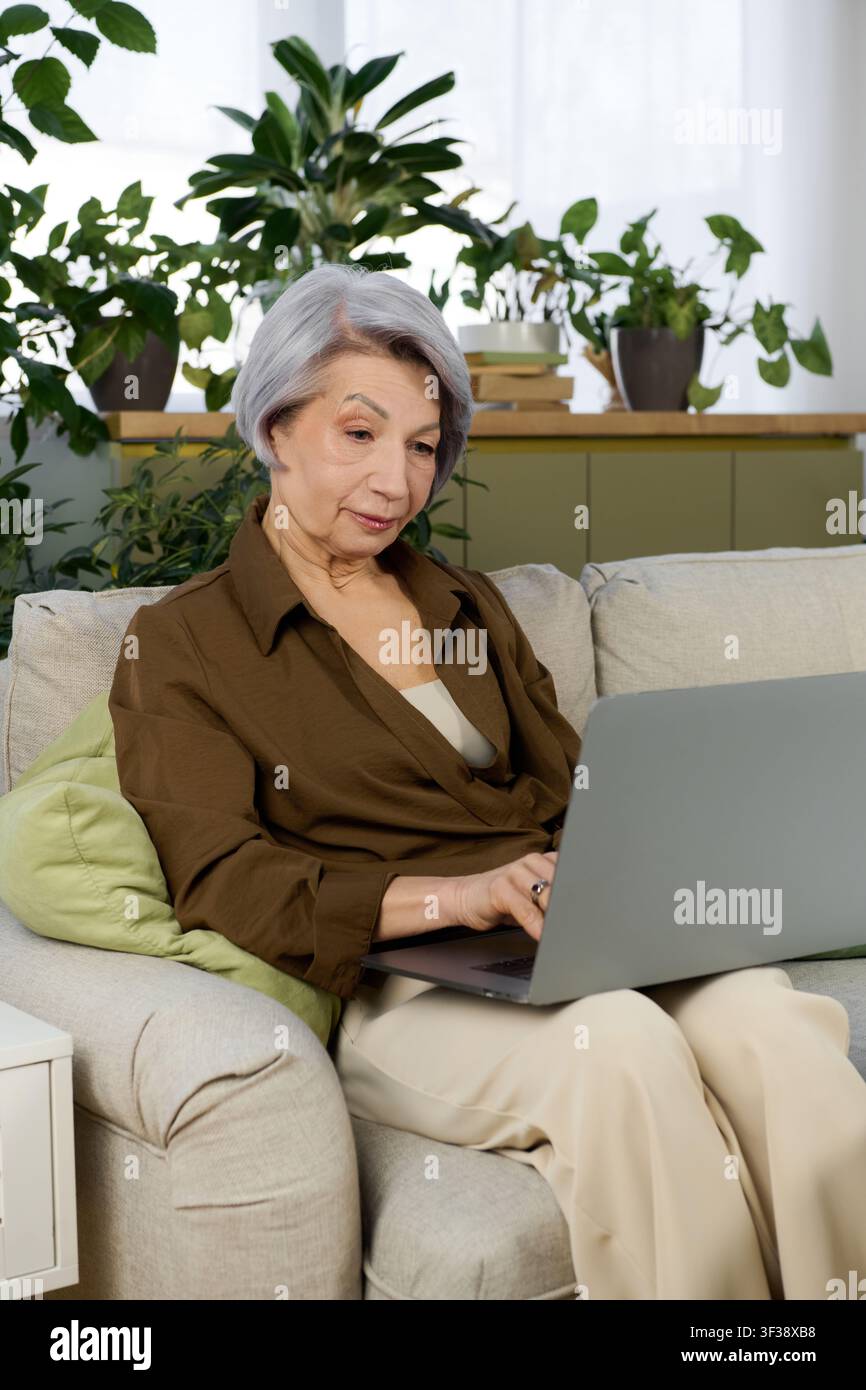 Una donna anziana dedicata si impegna con il suo laptop in un ufficio domestico pieno di verde Foto Stock