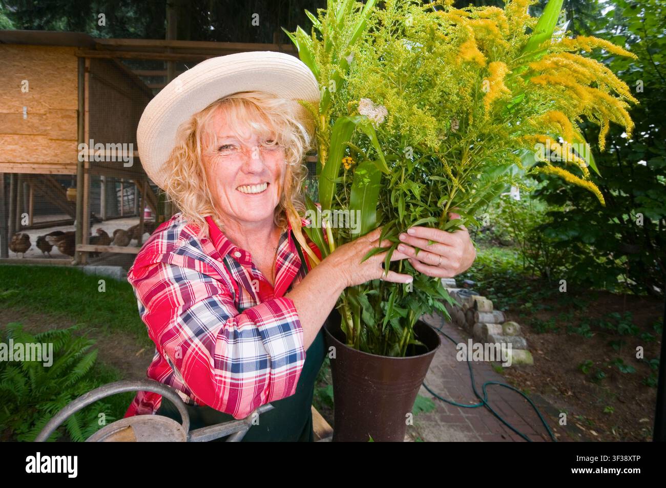 Ritratto dalla vita verso l'alto di una giardiniera bionda che indossa un cappello di paglia, sorride e tiene un mazzo di fiori verso la fotocamera. Foto Stock