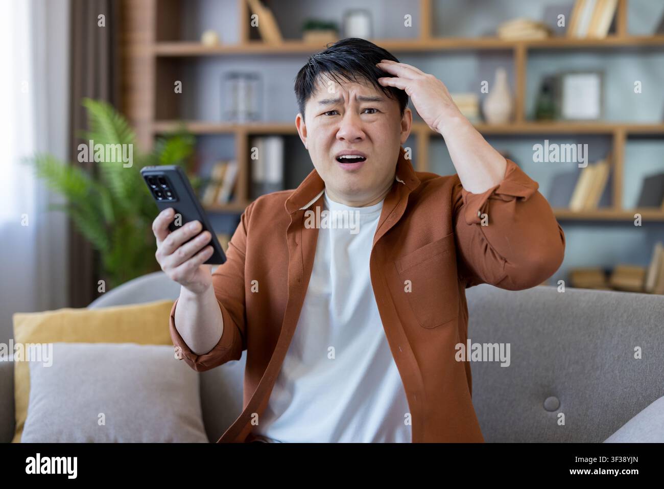 Uomo asiatico dall'aspetto confuso e stressato, che tiene uno smartphone mentre riceve notizie cattive scioccanti o sperimenta un errore digitale che causa frustrazione Foto Stock