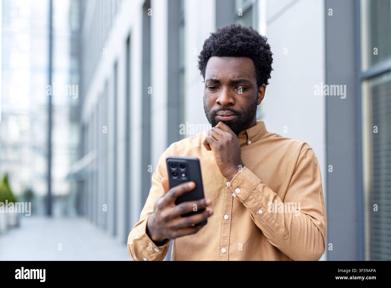 Giovane uomo d'affari in una camicia casual si trova fuori da un edificio per uffici, fissando il suo smartphone con un'espressione pensierosa e preoccupata mentre legge messaggi e dati Foto Stock