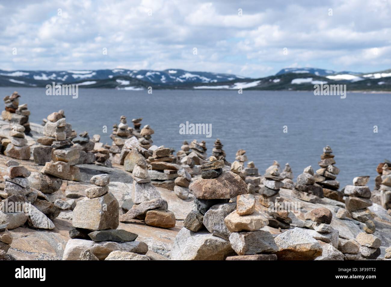 Innumerevoli cumuli di pietre impilati costeggiano un rivo roccioso sul Hardangervidda, in Norvegia, con montagne coperte di neve sotto un cielo nuvoloso. Foto Stock