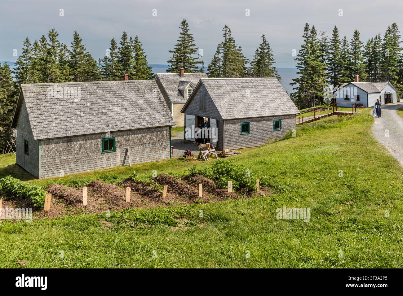 I visitatori camminano lungo un sentiero di ghiaia tra diversi edifici rivestiti di scandole al Highland Village Museum | Baile nan Gàidheal a Iona, Nuova Scozia, Canada, con vista sul lago Bras d'Or. Questa sezione del museo all'aperto presenta laboratori ed edifici residenziali risalenti al periodo compreso tra il 1880 e il 1920 Foto Stock