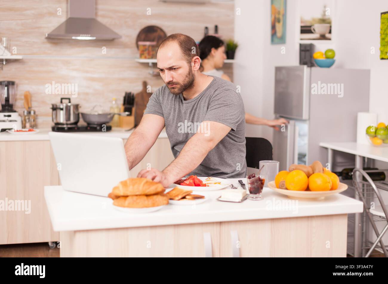Freelancer furioso urla al tavolo della cucina mentre sua moglie prepara la colazione, mostrando lo stress lavorativo e la tensione mattutina a casa per un uso realistico in stock commerciale e contesto specifico della fila. Foto Stock