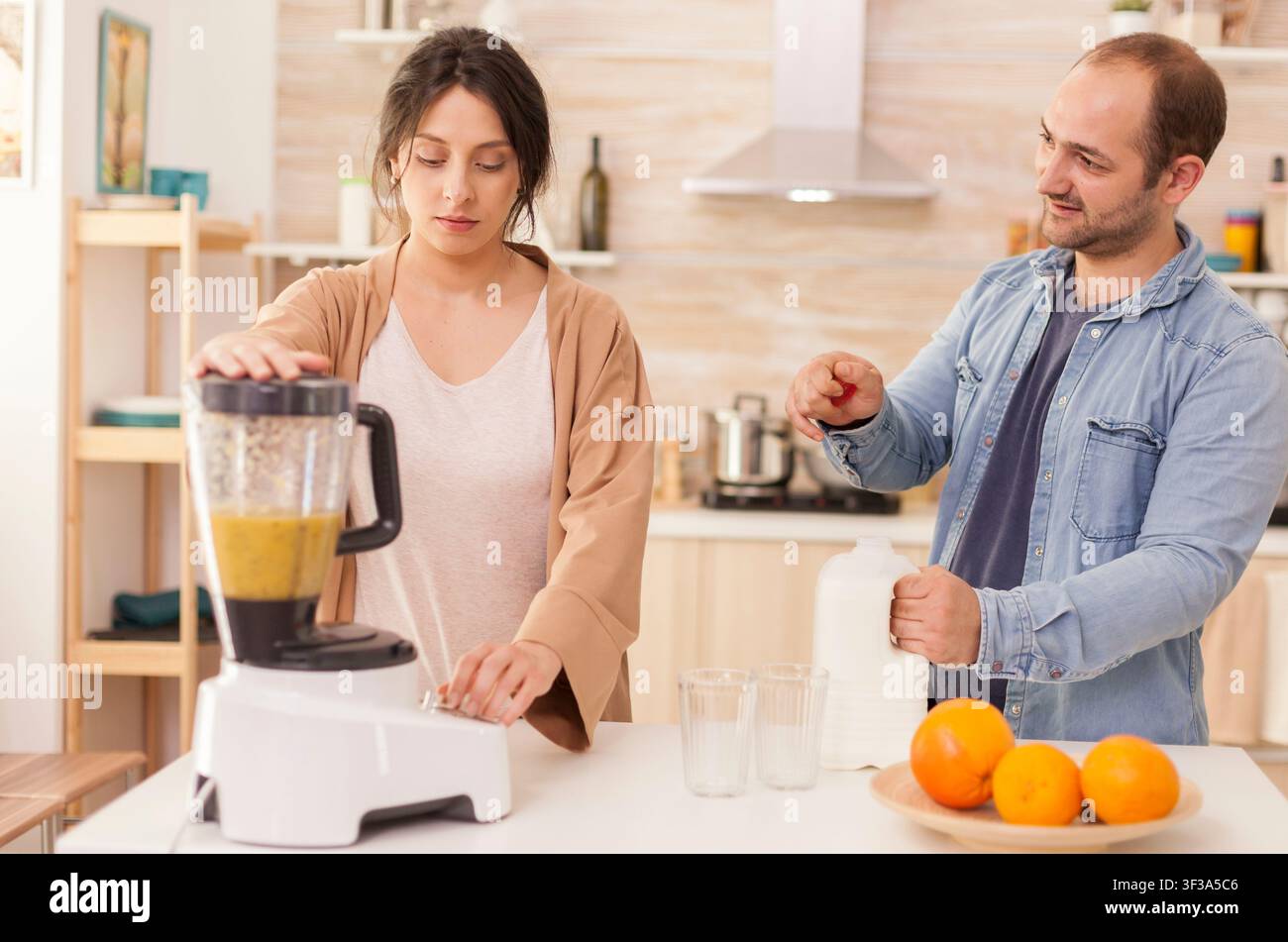 Donna mescola frutta in un frullatore per frullati mentre suo marito apre una bottiglia di latte, preparando insieme una colazione sana in cucina. Foto Stock