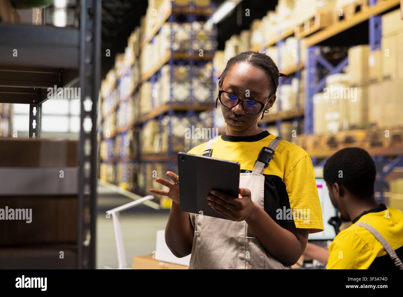 Impiegata industriale nera che lavora su attività di elaborazione degli ordini con tablet, il centro di distribuzione garantisce un funzionamento efficiente del magazzino. Gestione dei dati dei pacchi per la consegna e-commerce. Foto Stock