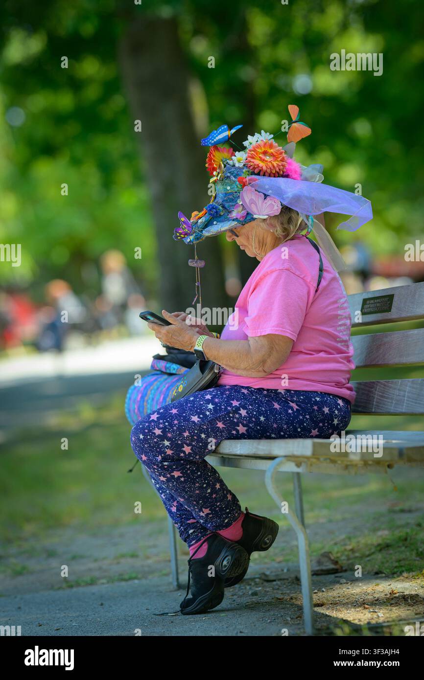 Donna seduta su una panchina del parco con un cappello ornato di fiori e decorato a un festival musicale Foto Stock