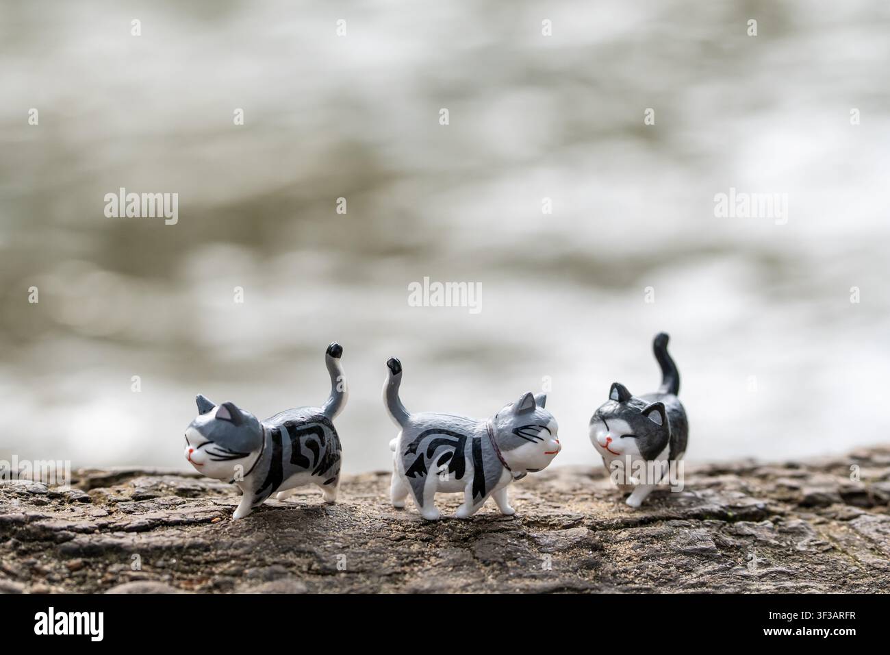 Chiang Mai, Thailandia - 13 luglio 2026: Tre figurine di gatti grigi e bianchi che camminano insieme con la coda sollevata su una radice d'albero accanto a una cascata Foto Stock