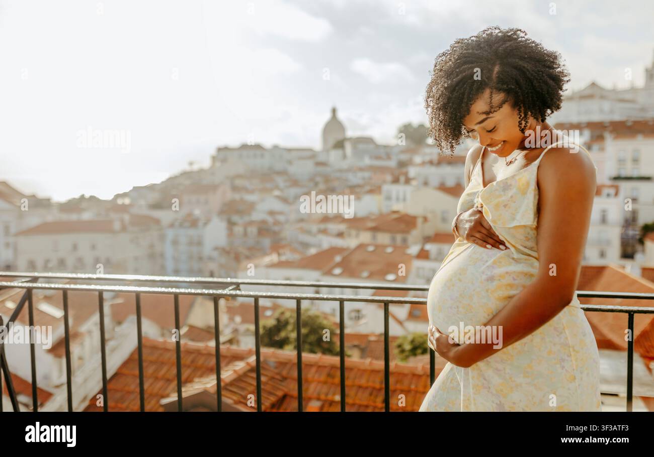La giovane donna afroamericana sorridente in abito tocca la pancia grande con le mani, cammina in città Foto Stock