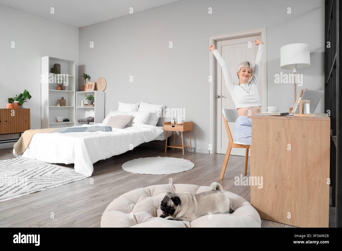 Giovane donna con cane carlino che si stiracchia al tavolo in camera da letto Foto Stock