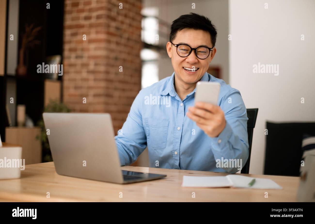 Imprenditore asiatico entusiasta che utilizza smartphone e laptop, lavora online seduto al posto di lavoro in un ufficio domestico Foto Stock