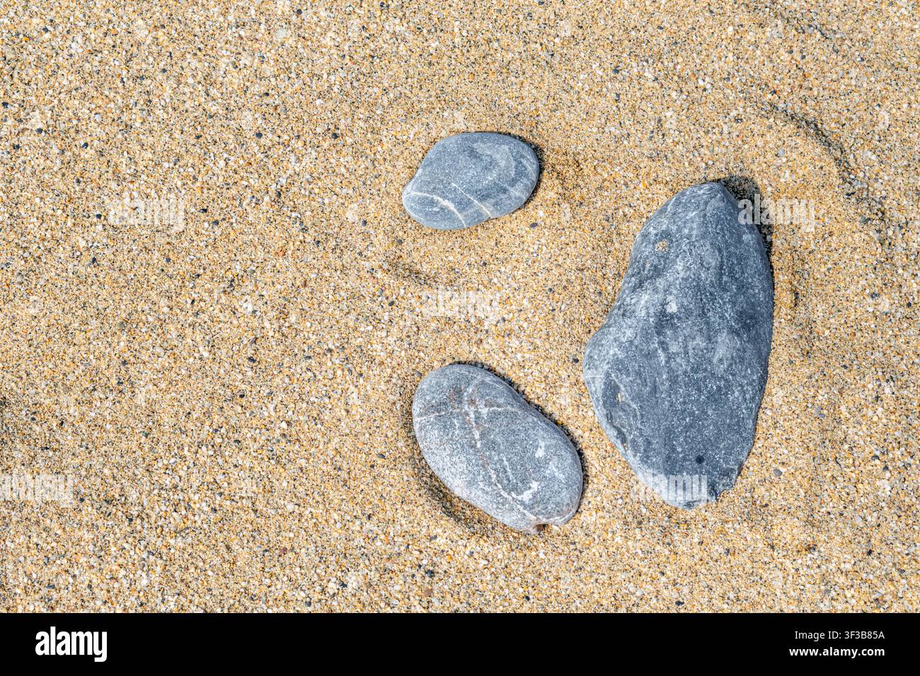Tre pietre grigie naturali distese su sabbia dorata della spiaggia. Composizione minimalista con texture organiche e luce solare. Sfondo versatile Foto Stock