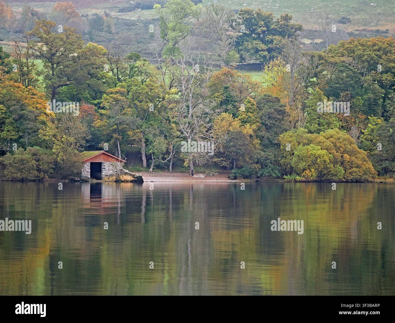 Vista del paesaggio autunnale sulle acque calme di Ullswater nel Lake District inglese con tipico capannone per barche in pietra sulla riva boscosa - Cumbria, Inghilterra, Regno Unito Foto Stock