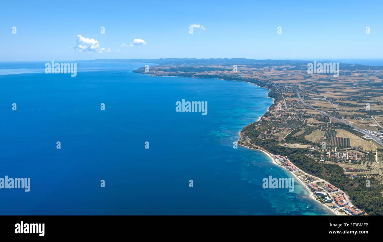 Vista aerea della costa di Kassandra in Grecia che mostra un mare blu chiaro, la linea costiera, campi agricoli e edifici sparsi sotto un cielo parzialmente nuvoloso Foto Stock