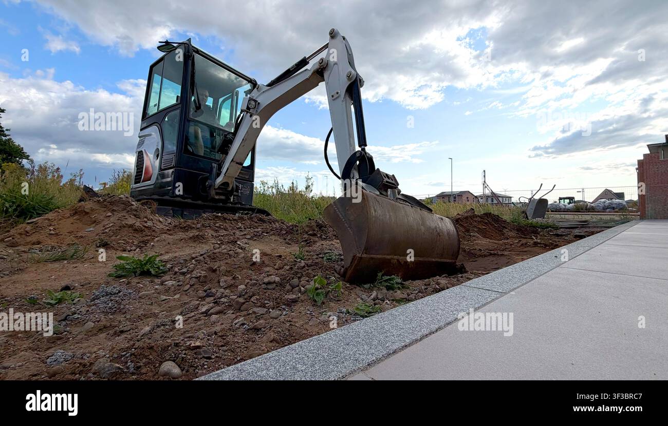 Operatore in escavatore che scava terra in un cantiere edile con vegetazione verde e cielo nuvoloso sullo sfondo Foto Stock