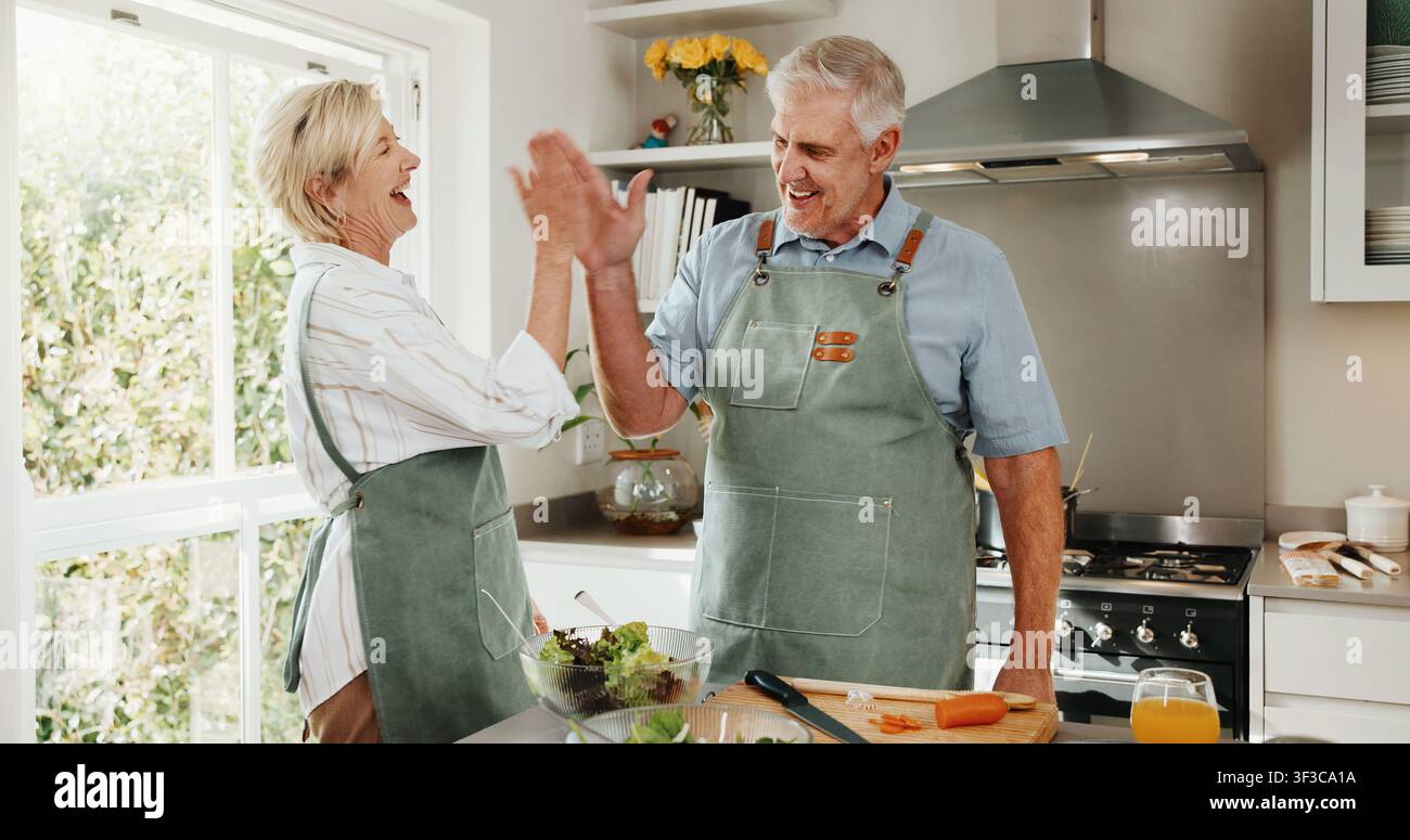 Anziano, coppia e cucina in cucina con high five, legame insieme e ridono per una battuta divertente. Persone anziane felici e sposate e celebrazione in Foto Stock
