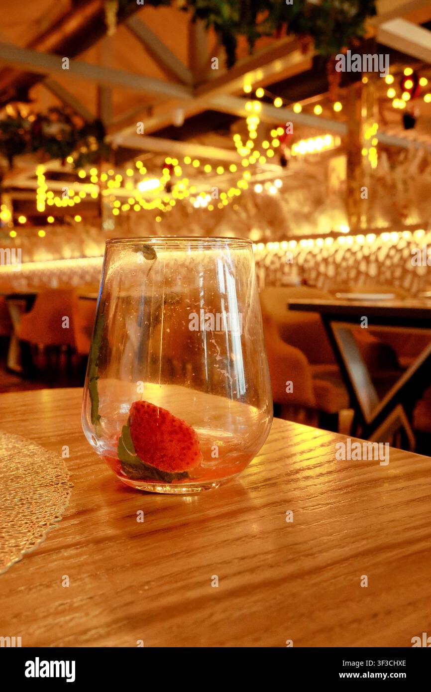 Bicchiere di vino vuoto con una fragola fresca rimasta dopo il drink su un tavolo di legno in un ristorante accogliente. Le luci calde della sera creano un'atmosfera romantica per la cena. Foto Stock