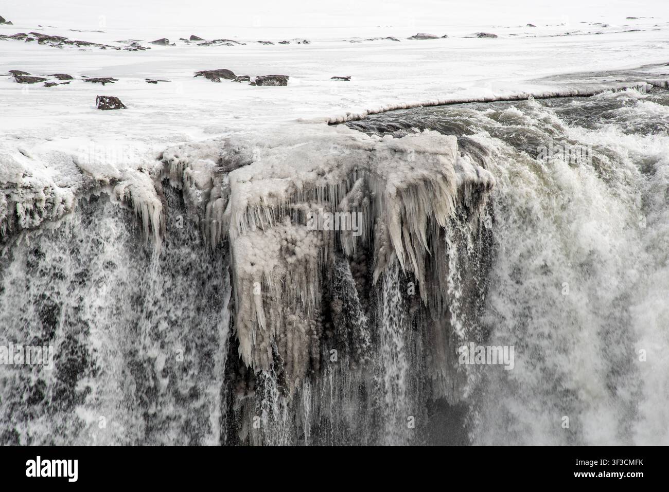 Dettaglio ad angolazione alta di acqua scura che si riversa oltre il bordo di una scogliera vulcanica. Blocchi di ghiaccio spessi e lunghe formazioni di ghiaccioli ricoprono la roccia, fondendosi con il Foto Stock