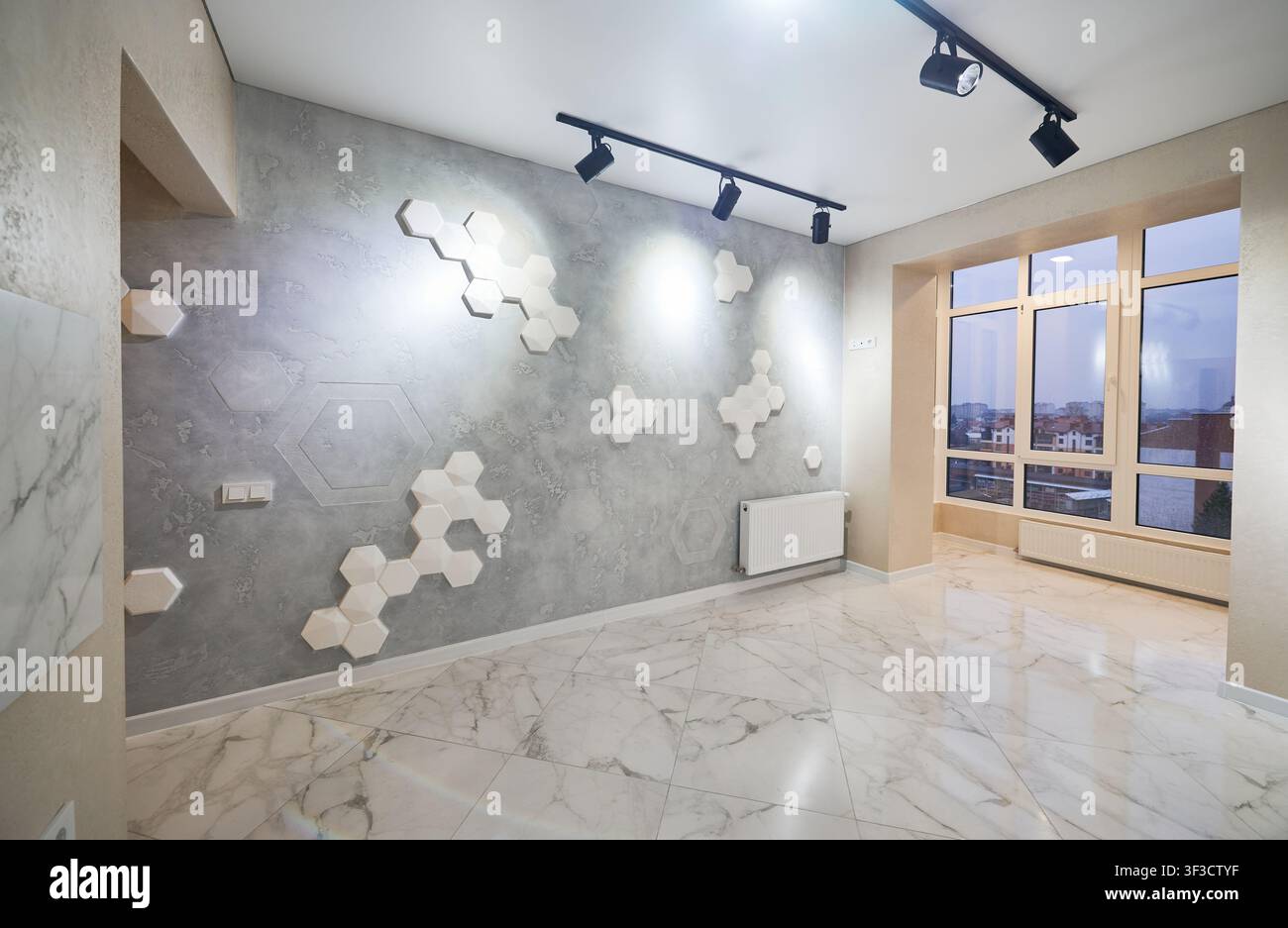 Stanza vuota moderna con pannelli murali esagonali decorativi, faretti neri su binario e grandi finestre, ampia vista sull'interior design elegante di un appartamento. Concetto di design contemporaneo e ristrutturazione Foto Stock