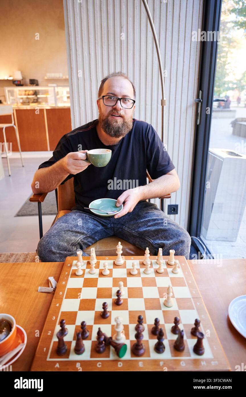 Uomo di fronte alla telecamera con scacchiera e tazza, espressione invitante e prontezza per una rivincita, ambientazione di gioco casuale con vista su finestra di vetro, pasticcino su piatto e luce ambientale calda, atmosfera conversazionale Foto Stock