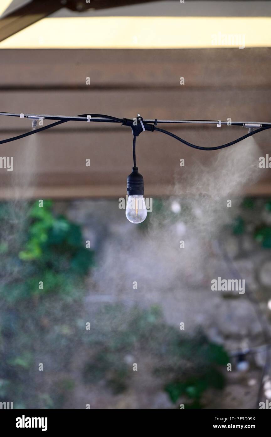 Primo piano di una linea di nebulizzazione esterna che spruzza una fine nebbia di vapore acqueo accanto a una lampadina Edison retrò nera su una terrazza di caffè. Progettata per rinfrescare le aree di seduta all'aperto durante le calde giornate estive, creando un'atmosfera confortevole e rinfrescante. Foto Stock