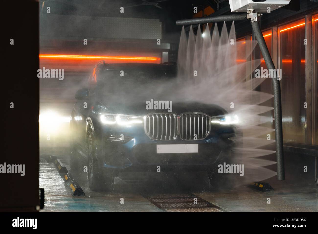 SUV premium blu si muove attraverso il lavaggio automatico, getti d'acqua che colpiscono la carrozzeria, nebbia che riempie il corridoio, strisce neon arancioni Foto Stock
