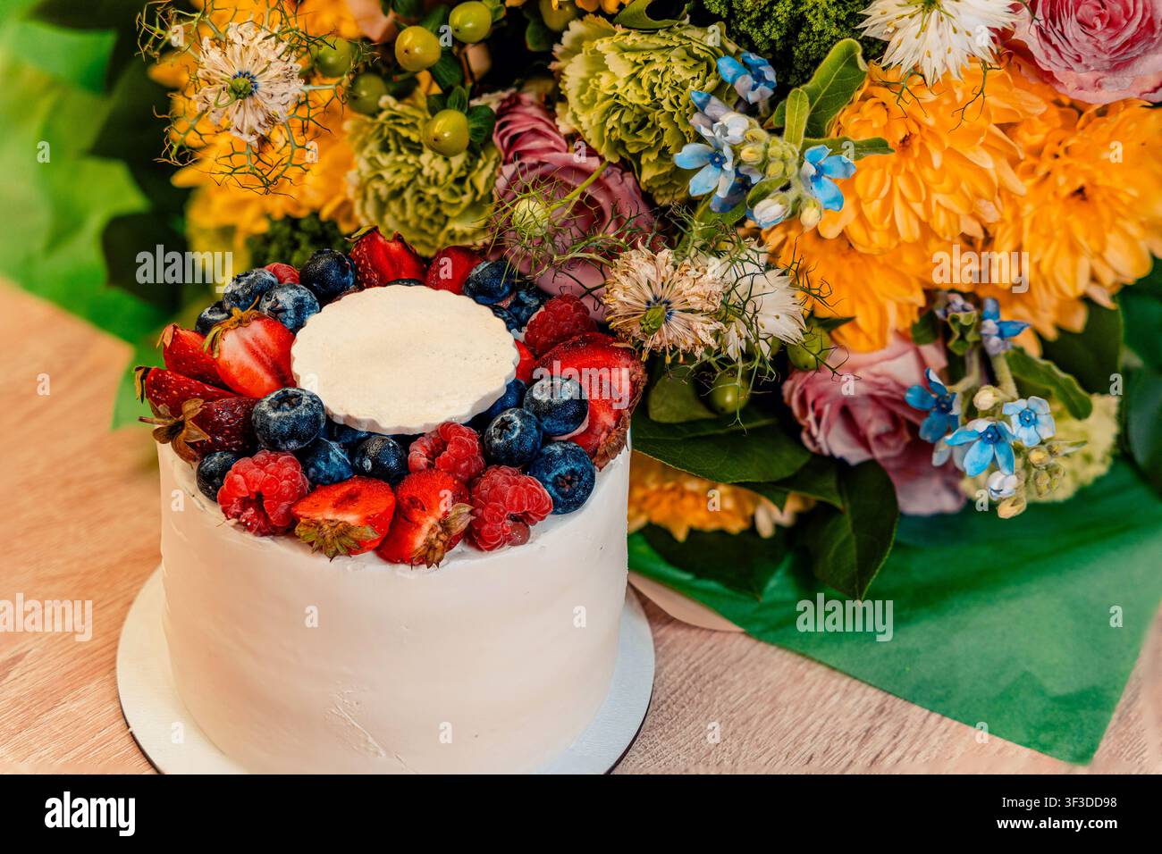Una torta bianca rotonda ricoperta di bacche è affiancata da cupcake abbinati e fiori vivaci, con toni caldi sul tavolo Foto Stock