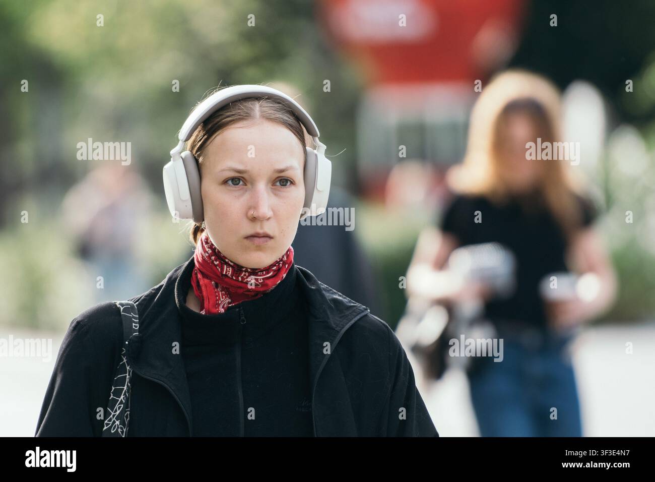 Bella ragazza che cammina con le cuffie e indossa una giacca casual nera con cerniera abbinata a un vivace foulard Paisley rosso legato ordinatamente intorno al collo Foto Stock