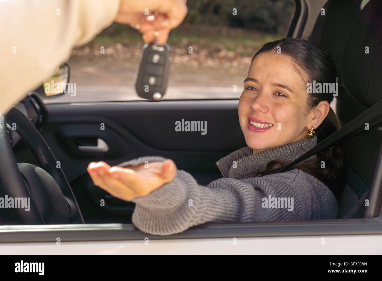 Giovane donna sorridente che guarda la chiave dell'auto che le viene consegnata attraverso il finestrino, interno dell'auto, concetto di successo Foto Stock