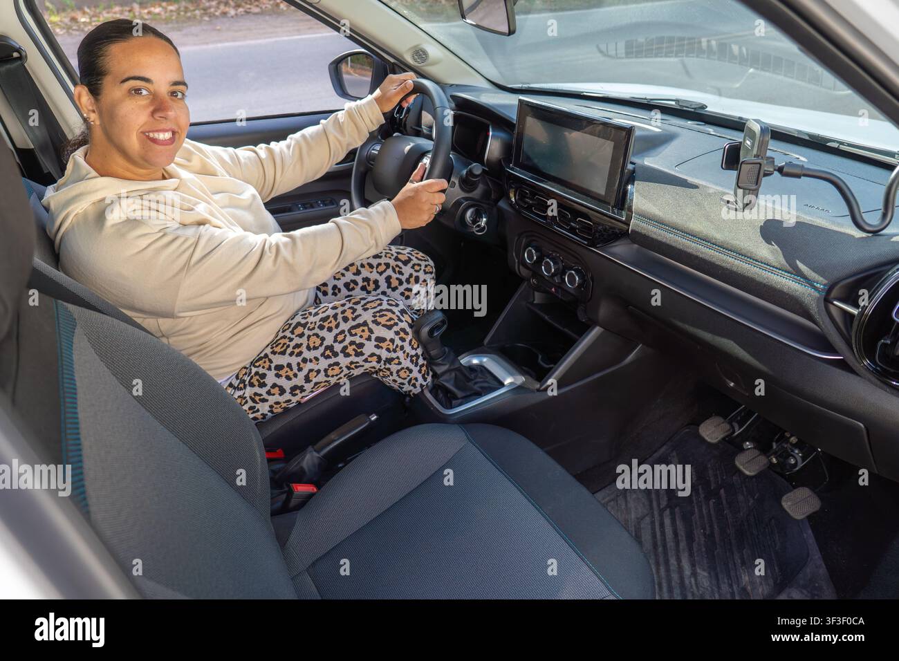 Donna sul sedile del conducente sorride e guarda verso la fotocamera, angolazione dall'alto, interno dell'auto con cruscotto e supporto per telefono visibile Foto Stock