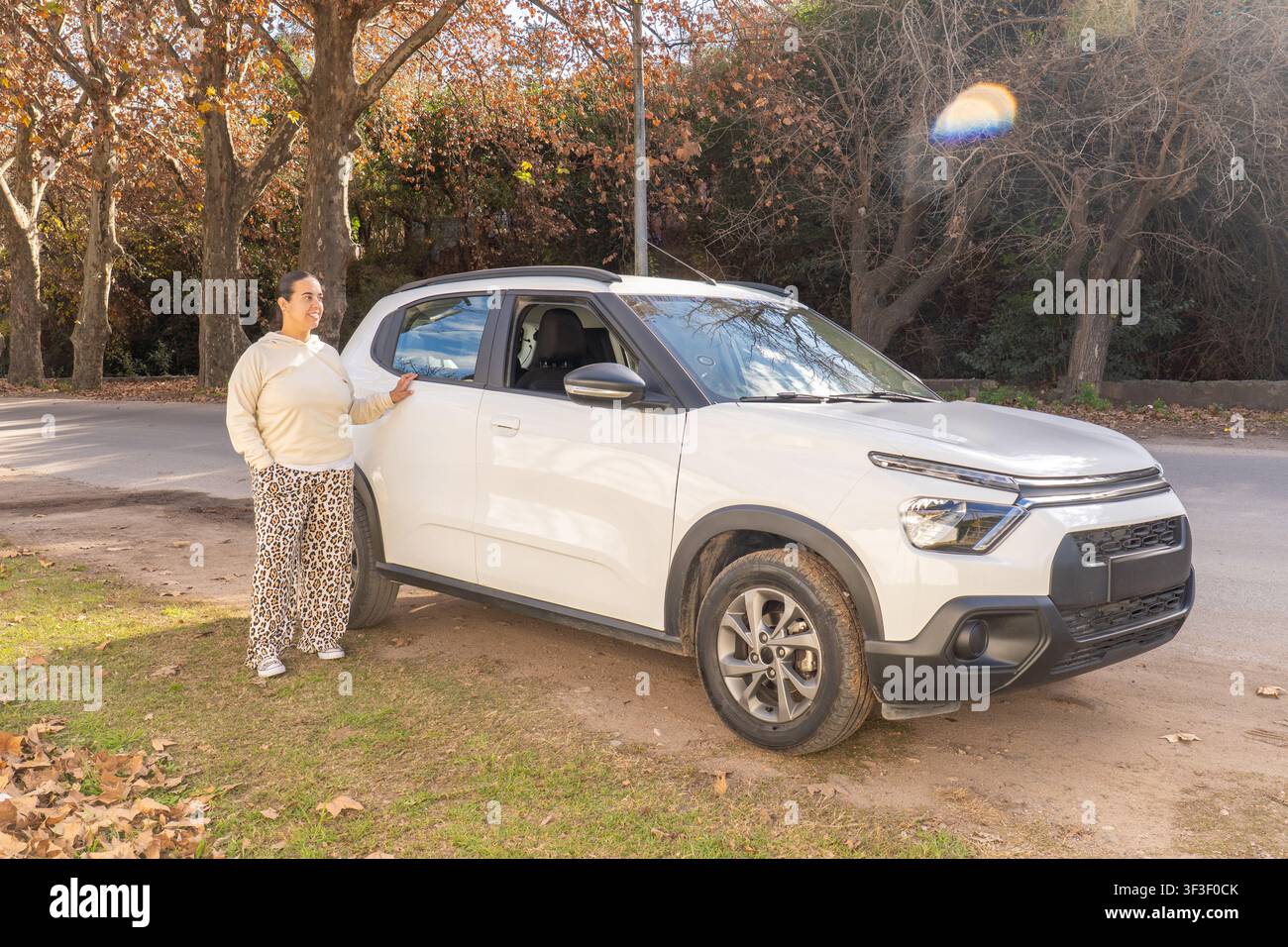 Donna in piedi accanto a un SUV bianco con la mano sulla portiera, che guarda all'indietro sopra la spalla, veduta a tutto corpo in ambiente autunnale all'aperto Foto Stock