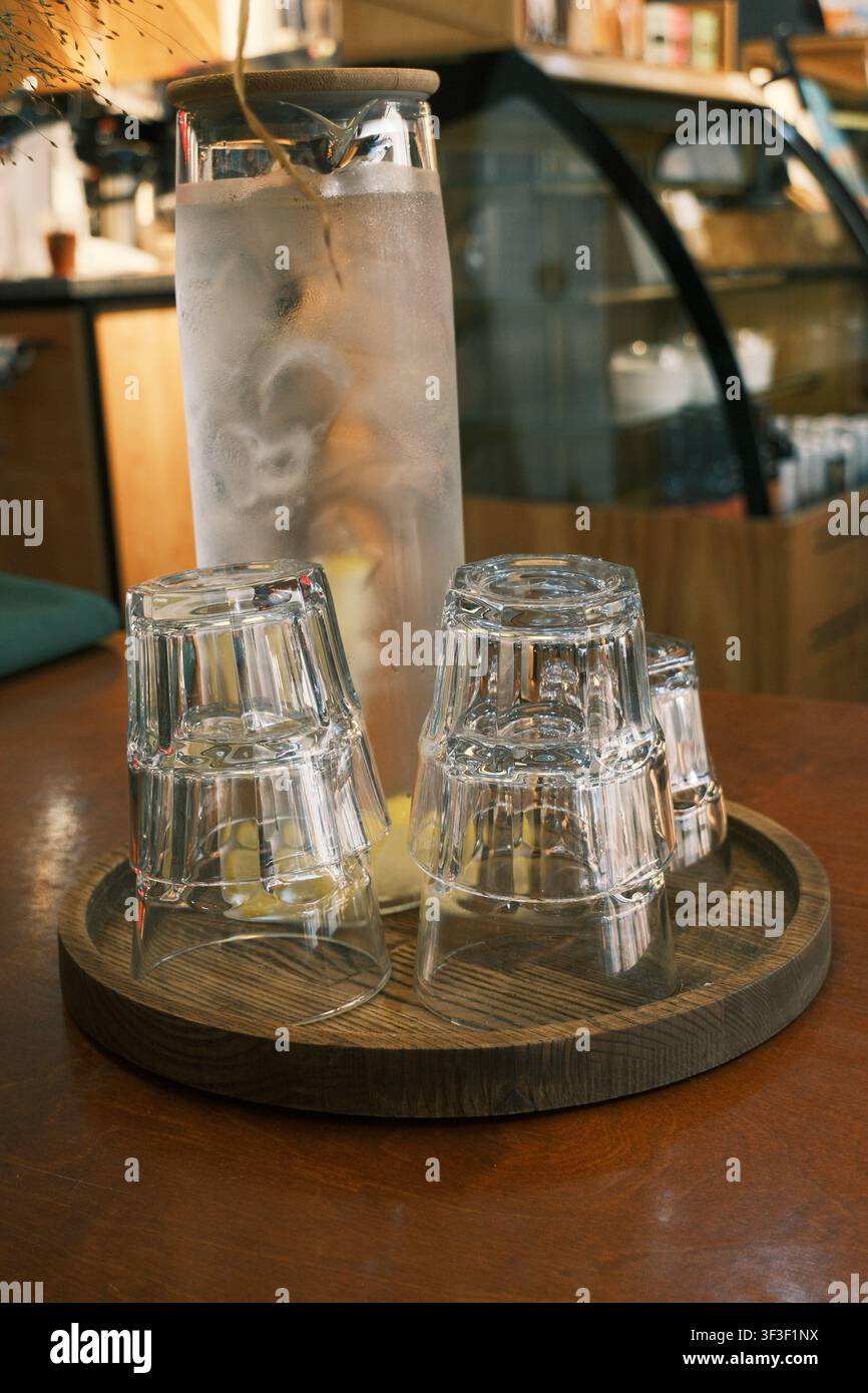 Primo piano di una brocca di vetro ghiacciata con acqua al limone ghiacciata affiancata da bicchieri trasparenti impilati su un vassoio di legno rustico in un ambiente accogliente e caldo di un caffè Foto Stock