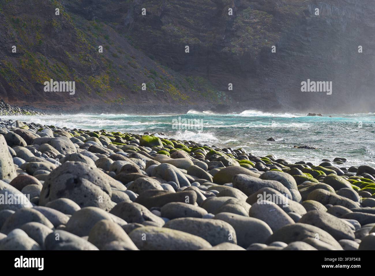 Un'onda turchese che si infrange verso la spiaggia di ciottoli ricoperta di muschio di Playa del Fraile sotto le scogliere nebbiose Foto Stock