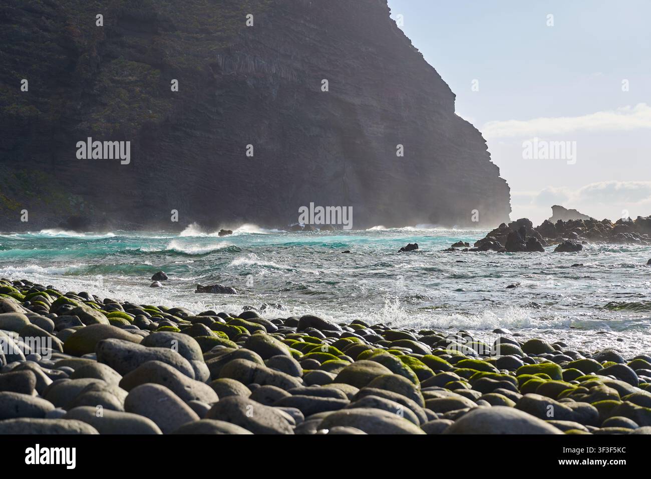 Un'onda turchese che si infrange verso la spiaggia di ciottoli ricoperta di muschio di Playa del Fraile sotto le scogliere nebbiose Foto Stock