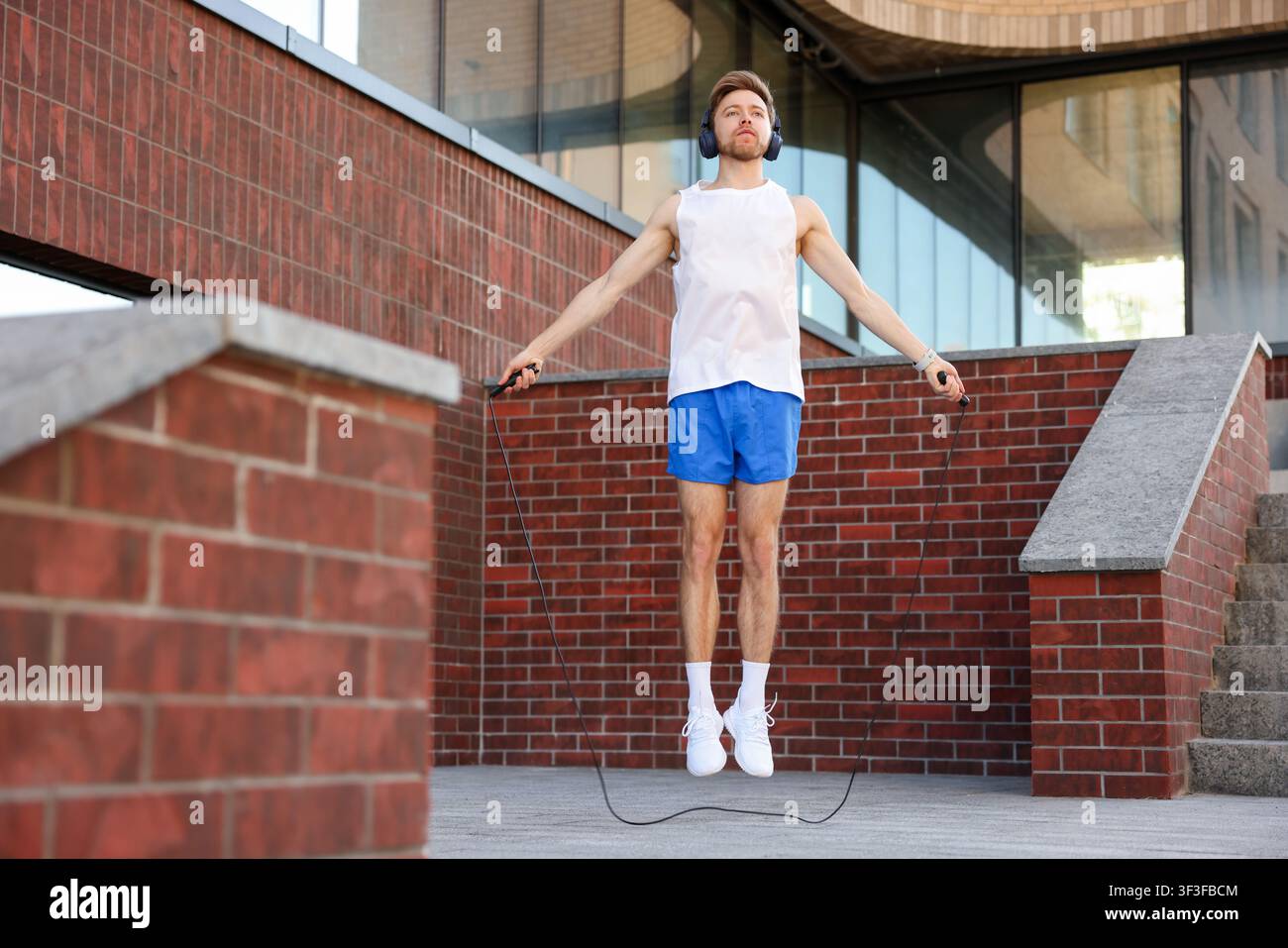 Uomo in abbigliamento sportivo e cuffie che fa esercizio con la corda per saltare vicino a un edificio all'aperto, vista dall'alto Foto Stock